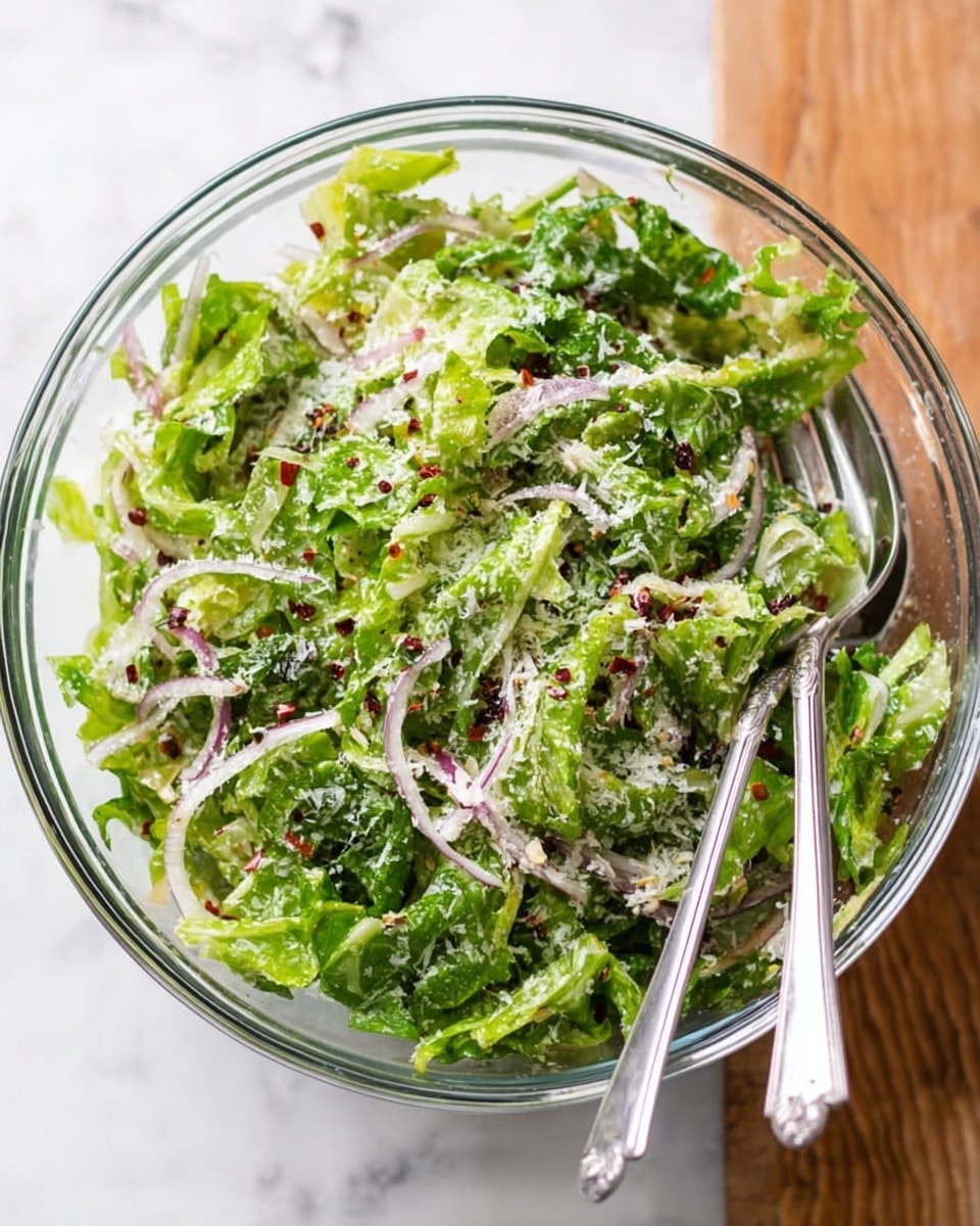 A clear glass bowl filled with a fresh green salad made of torn lettuce leaves and thin, curved slices of light purple onion scattered evenly on top. The salad is sprinkled with white grated cheese and tiny red chili flakes, adding color and texture. Two silver forks rest inside the bowl, one standing upright and the other lying on the side. The bowl sits on a white marbled surface. photo taken with an iphone --ar 4:5 --v 7