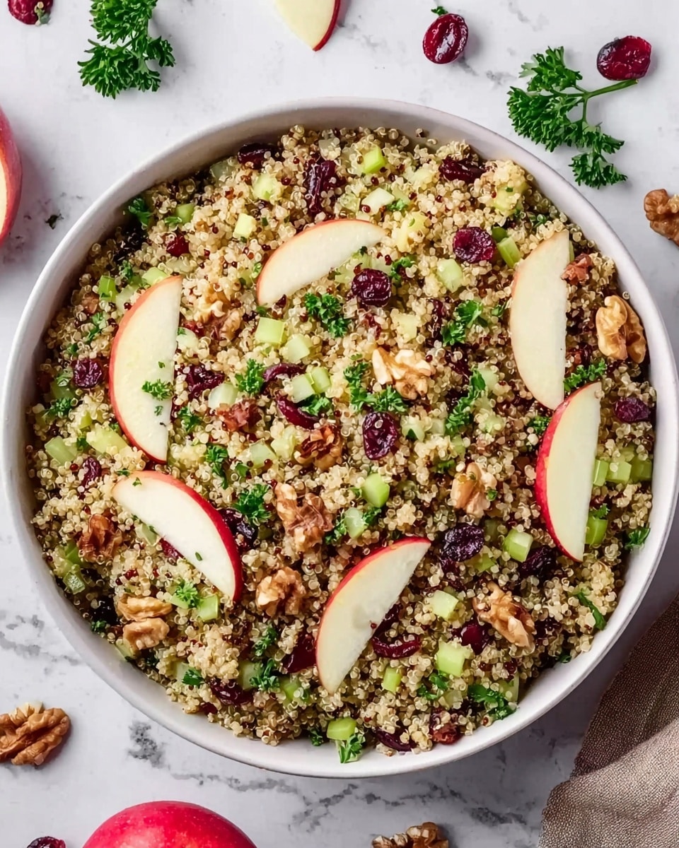 A white bowl filled with a mixed quinoa salad showing a base layer of fluffy, pale yellow quinoa grains spread evenly. On top, there are small pieces of green celery and green parsley leaves adding fresh color, along with chopped red and white apple slices scattered across the salad. Mixed in are dark red dried cranberries and brown walnut pieces placed throughout, giving the dish a varied and textured look. The bowl sits on a white marbled surface with some apple slices and dried cranberries placed nearby as decoration. Photo taken with an iphone --ar 4:5 --v 7