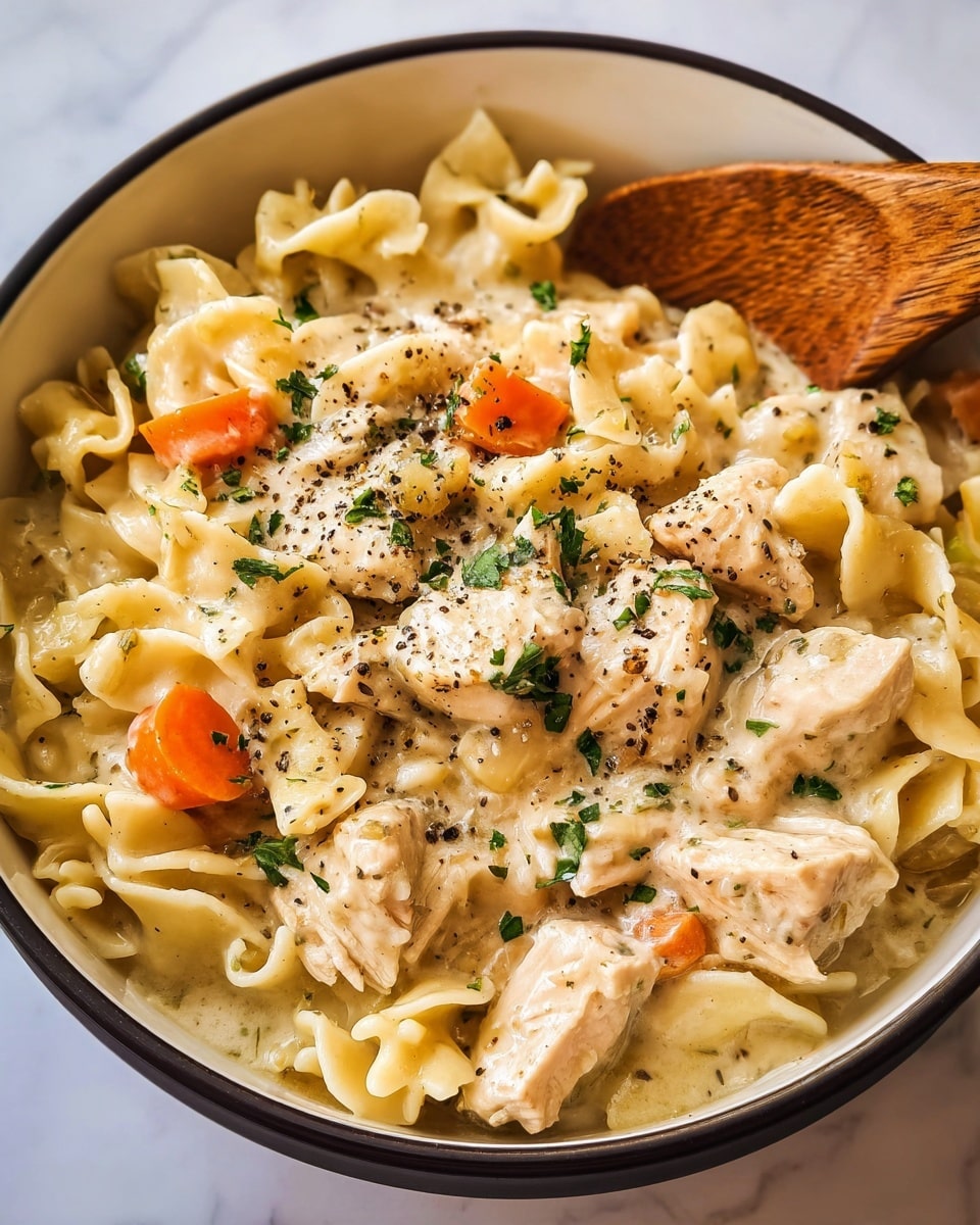 A close-up view of a creamy chicken noodle dish served in a white bowl with a dark rim. The dish has three main layers: the bottom layer is soft, wavy egg noodles coated in a light beige creamy sauce; the middle layer includes chunks of cooked chicken breast in a pale white color and small pieces of bright orange carrot; the top layer is sprinkled with freshly chopped green herbs and cracked black pepper, adding texture and color contrast. A wooden spoon is partially seen resting on the right side of the bowl. The bowl sits on a white marbled surface. photo taken with an iphone --ar 4:5 --v 7