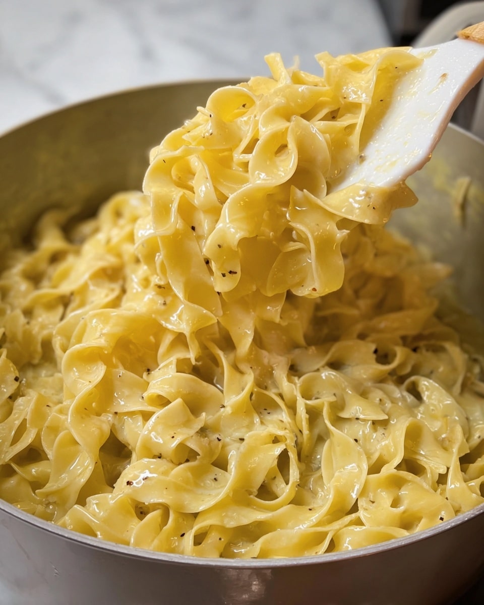 A close-up view of creamy cooked egg noodles sits in a pot, with a white spatula lifting a portion to show its soft, glossy texture. The noodles are thick, yellowish with a slight sheen from the creamy sauce coating them, and have a twisted, ribbon-like shape with some specks of black pepper visible. The pot's interior is light gray, and the background has a subtle, blurred white marbled texture. The image focuses on the rich, smooth noodles glistening under soft light, highlighting their tenderness and creamy finish. photo taken with an iphone --ar 4:5 --v 7