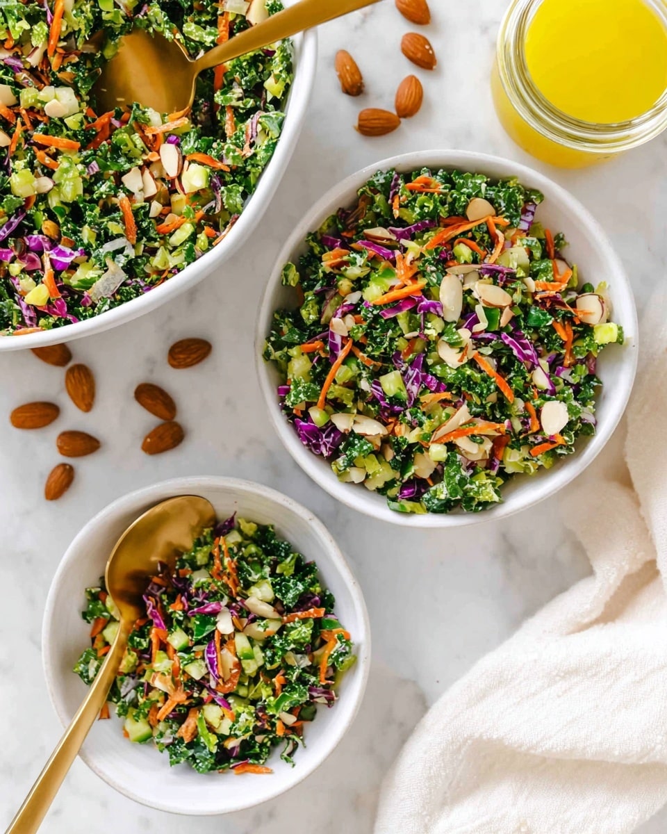 A large white bowl filled with a colorful chopped salad made of finely cut green kale, orange carrot shreds, small pieces of purple cabbage, and bits of cucumber, mixed with sunflower seeds and almonds scattered on top and around the bowl. A gold spoon rests inside the bowl on the right side, partially submerged in the salad. Surrounding the bowl on a white marbled surface are a small white bowl of sunflower seeds, a glass jar of yellow dressing, several whole almonds, and two lemon wedges near the top right. A light beige cloth is placed at the bottom right corner. Photo taken with an iphone --ar 4:5 --v 7