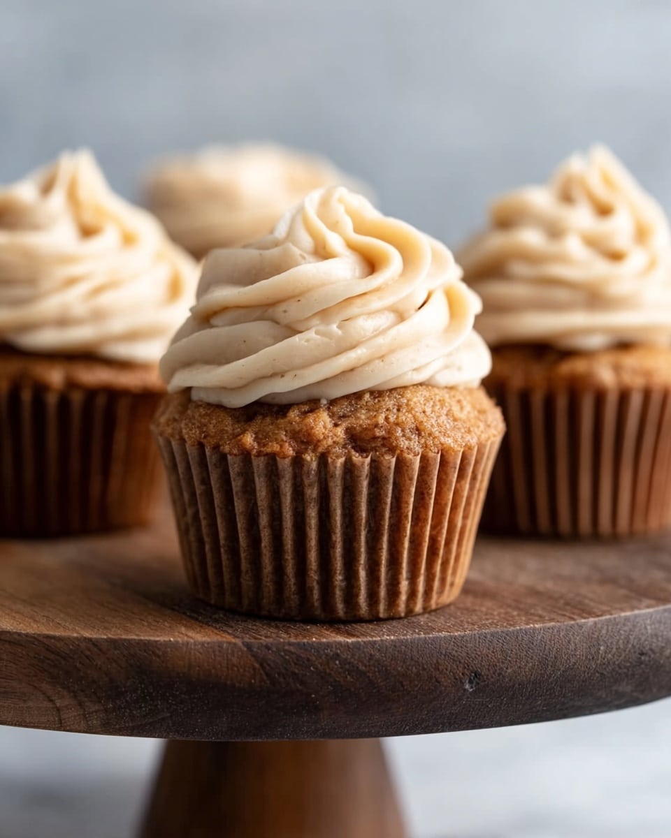 A close-up view of three cupcakes placed on a dark wooden cake stand against a white marbled background. Each cupcake has one layer of light brown cake with a slightly rough texture and is topped with a generous swirl of creamy beige frosting that is smooth and fluffy. The cupcakes have pleated brown wrappers, adding to the textured look. The scene focuses mainly on the middle cupcake, showing detailed swirls of frosting and the cake’s soft crumb, while the other two cupcakes are slightly blurred in the background. Photo taken with an iphone --ar 4:5 --v 7