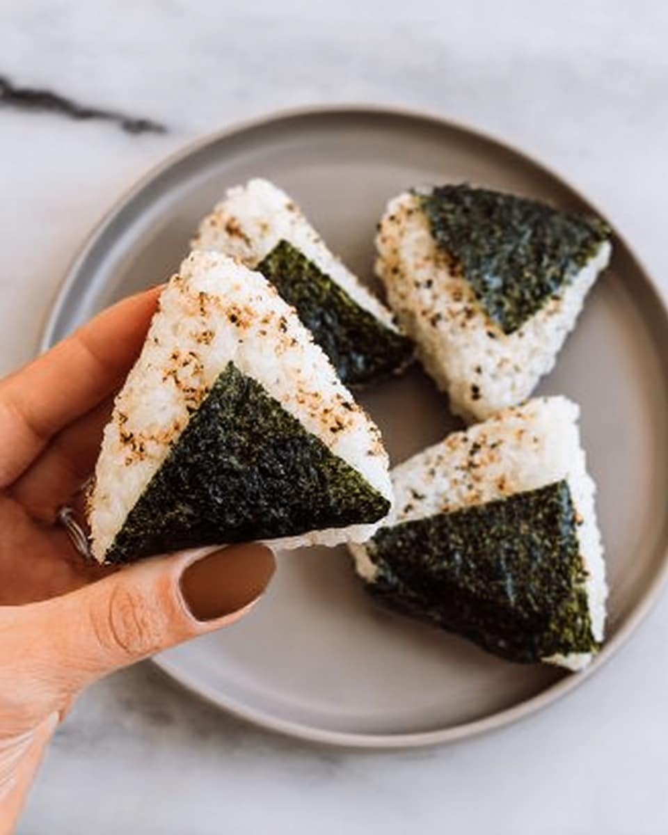 Three onigiri rice balls shaped like triangles are arranged in a row on a white plate with a blue floral rim. Each rice ball has a smooth, white rice dome in the center, wrapped at the base and partly up the sides with a dark green, shiny seaweed sheet. At the top of each rice ball, there is a generous scoop of light brown shredded tuna salad with a creamy texture. The scene is set on a white marbled surface with small dishes and containers blurred softly in the background. Photo taken with an iphone --ar 4:5 --v 7