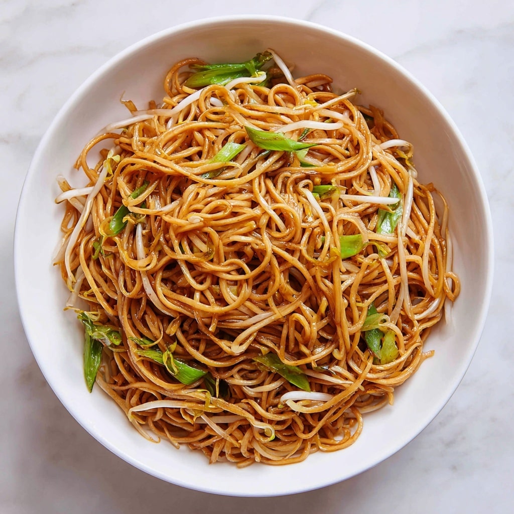 A white bowl is filled with a large serving of thin, cooked noodles that have a rich brown color from the sauce. Mixed throughout the noodles are light beige bean sprouts and bright green pieces of chopped green vegetables, giving some contrast to the dish. The noodles look slightly glossy and tangled together, showing a textured surface. The bowl sits on a white marbled surface with a soft, clean background, which enhances the warm tones of the noodles and greens. Photo taken with an iphone --ar 4:5 --v 7
