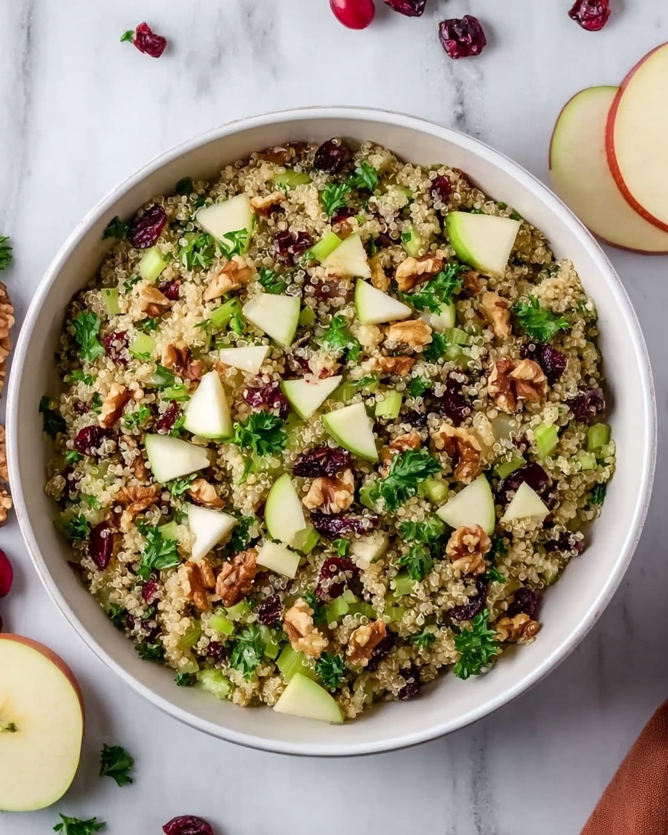 A large white bowl is filled with a layered quinoa salad, the base layer consisting of small, round, beige and slightly translucent quinoa grains. Mixed throughout are small, bright green celery pieces adding a crunchy texture. Scattered among these are thin, curved slices of red-skinned apple with white flesh, offering a fresh and crisp look. Deep red dried cranberries are dotted evenly across the salad, along with light brown walnut halves adding a rough, textured appearance. Small dark green parsley leaves are sprinkled on top, creating a fresh and colorful contrast. The bowl rests on a white marbled surface, with some apple halves, cranberries, and parsley around, enhancing the freshness of the scene. Photo taken with an iphone --ar 4:5 --v 7