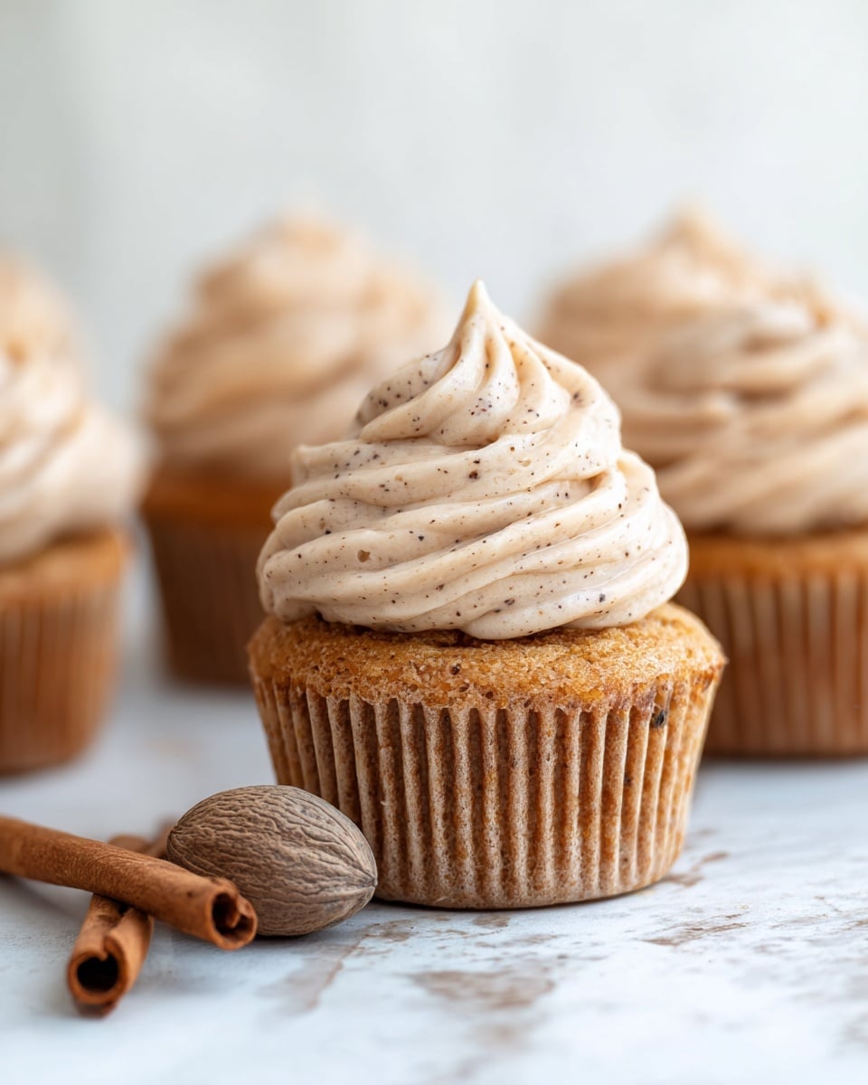 A close-up view of several brown cupcakes showing one in the front with a thick swirl of light beige frosting speckled with tiny dark dots, creating a smooth and creamy texture on top. The cupcake’s bottom layer is a golden-brown cake with a slightly rough texture and visible paper liner ridges. In front of the cupcake, there is a whole nutmeg seed and a cinnamon stick lying on a white marbled surface. More cupcakes with the same frosting and cake layers are blurred in the background. photo taken with an iphone --ar 4:5 --v 7