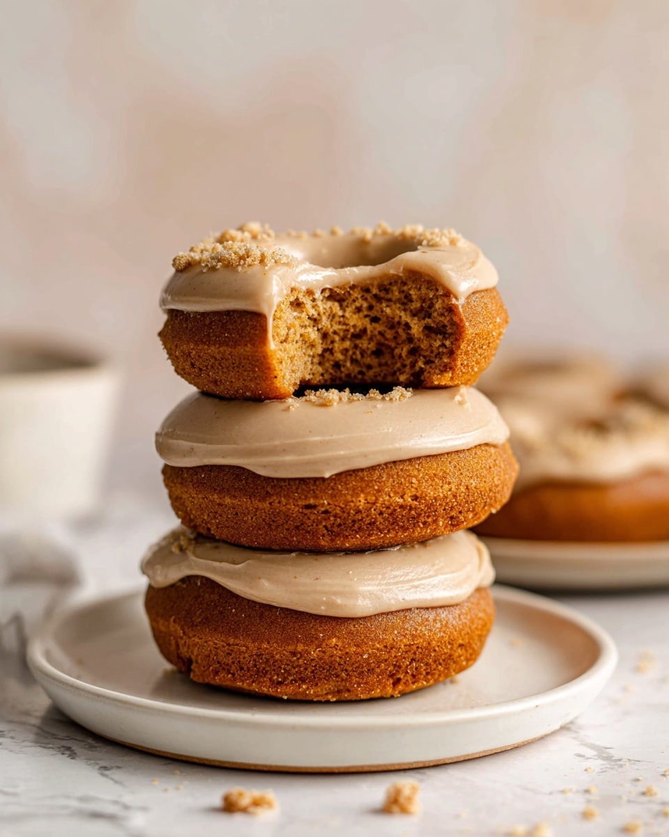 The image shows a stack of three round doughnuts on a white plate against a white marbled background. Each doughnut has a thick, smooth layer of creamy, light beige frosting on top, with small crumbles sprinkled over the middle doughnut's frosting. The doughnuts have a rich golden-brown color with a soft, spongy texture. The top doughnut has a bite taken out, revealing the moist and dense inside. Next to the plate, there is a half-eaten doughnut with the same frosting and color. The scene is softly lit, highlighting the textures and warm tones of the doughnuts. photo taken with an iphone --ar 4:5 --v 7