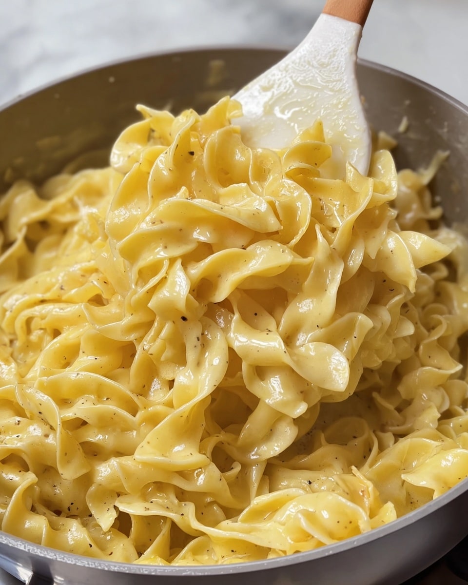 The image shows a close-up of creamy cooked egg noodles held above a white pot by a wooden-handled white spoon. The noodles are light yellow with a glossy, soft texture, slightly thick and curly. Small black specks of pepper are visible on the noodles, giving a subtle contrast to the creamy sauce covering them. The background is a white marbled texture. photo taken with an iphone --ar 4:5 --v 7