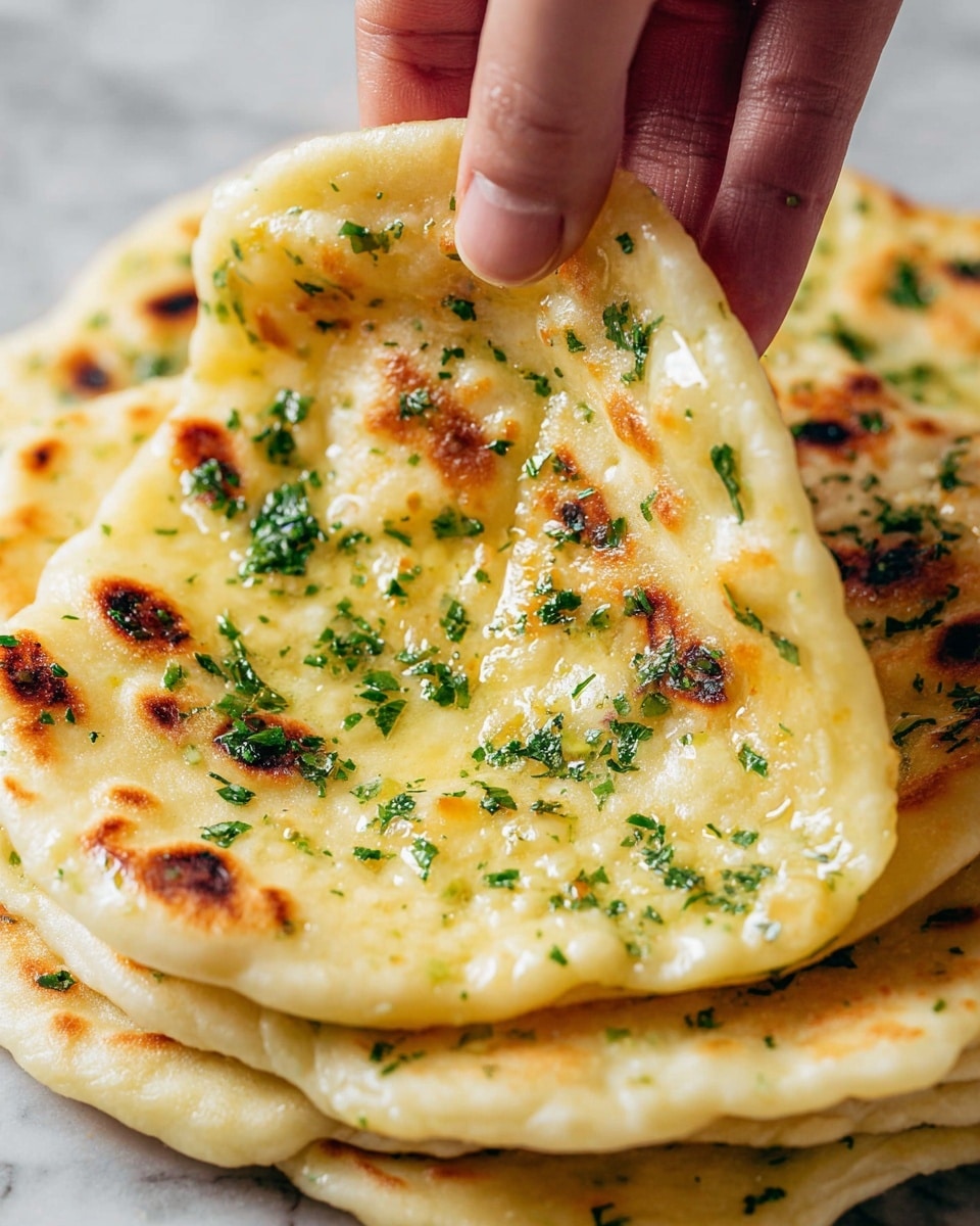 A close-up shot shows a stack of flatbreads, about three layers tall, each layer golden yellow with crisp, brown cooked spots and sprinkled with finely chopped green herbs. The texture appears soft and slightly oily, with some light bubbling on the surface. A woman's hand is lifting the top flatbread, bending it gently to show its thin, flexible texture. The background is a white marbled texture. Photo taken with an iphone --ar 4:5 --v 7