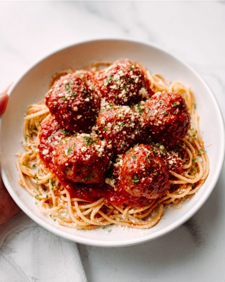A white oval plate filled with light brown cooked spaghetti noodles at the base, topped with a rich red tomato sauce. On top of the spaghetti, there are five large, round meatballs covered in the same red sauce, sprinkled with grated white cheese and small bits of green herbs. The plate is placed on a white marbled surface with a soft cloth partially visible. Photo taken with an iphone --ar 4:5 --v 7