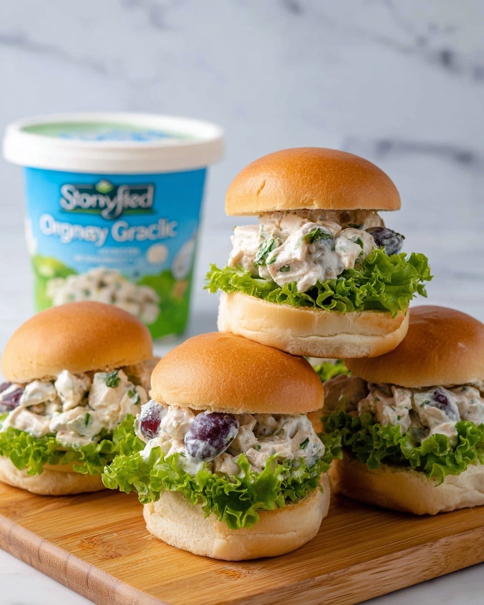The image shows four small sandwiches on a wooden board against a white marbled background. Each sandwich has two soft golden brown buns as the top and bottom layers. Inside, there is a layer of fresh green lettuce with a leafy texture, topped with a creamy salad filling that includes white chunks of chicken or tofu mixed with small dark purple pieces, possibly grapes or olives, and finely chopped green herbs. To the left, there is a white container of Stonyfield Organic Greek yogurt with a blue and green label, slightly blurred in the background. The sandwiches are arranged with two stacked vertically on the right side, and two placed next to each other on the left, showing the filling clearly. photo taken with an iphone --ar 4:5 --v 7