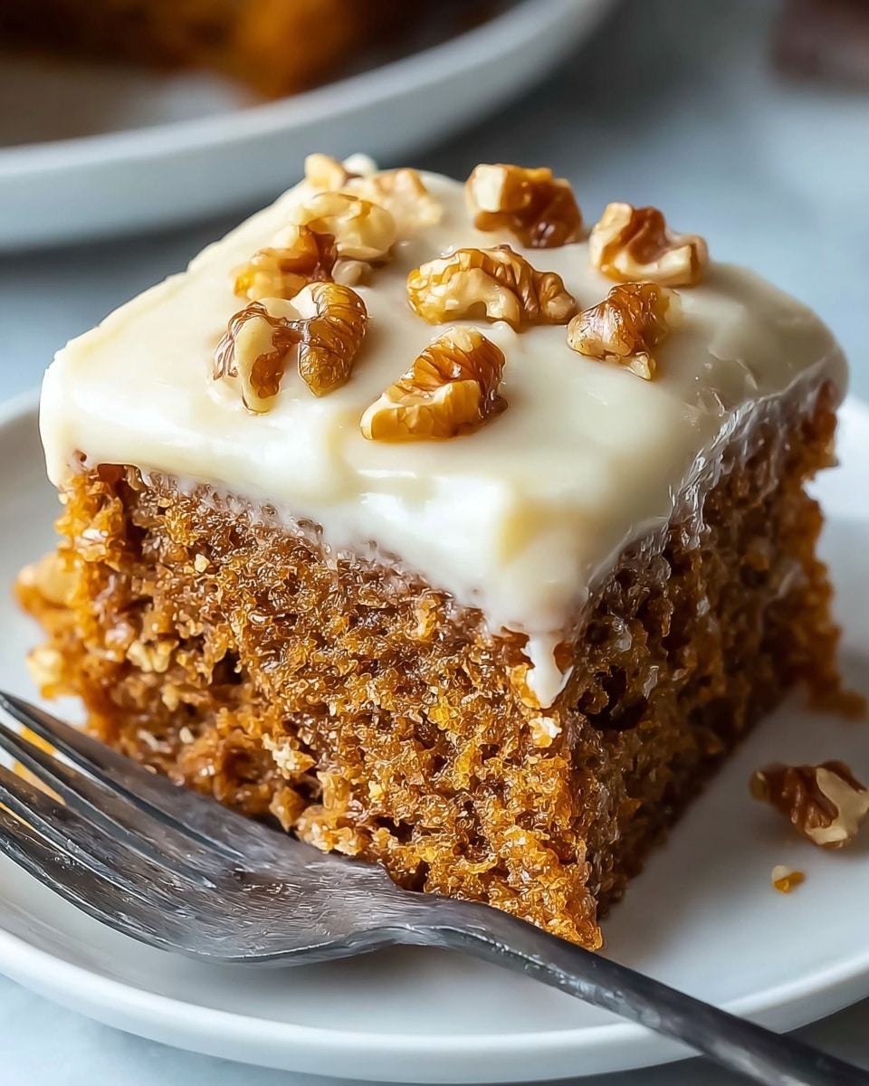 A close-up image shows a square piece of moist, light brown cake with a soft, crumbly texture. The cake has one thick layer topped with a smooth, creamy light beige frosting that slightly drips down the sides. On top of the frosting, there are several small, uneven pieces of chopped nuts with a mix of light and dark brown colors. The cake sits on a simple white plate with a silver fork next to it, all placed on a white marbled surface. Photo taken with an iphone --ar 4:5 --v 7