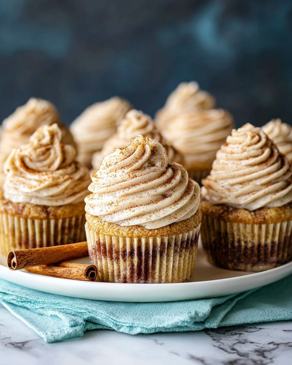A close-up of a cupcake with a bite taken out of it, showing a moist, speckled light brown cake base with a swirl of creamy, light beige frosting dusted with cinnamon on top. The cupcake liner is opened and placed on a white plate, with more cupcakes in the background, each topped with the same swirled frosting. The plate is set on a textured white marbled surface with a blue cloth partially visible beneath. The overall colors are warm, with soft lighting highlighting the texture of the frosting and cake. photo taken with an iphone --ar 4:5 --v 7