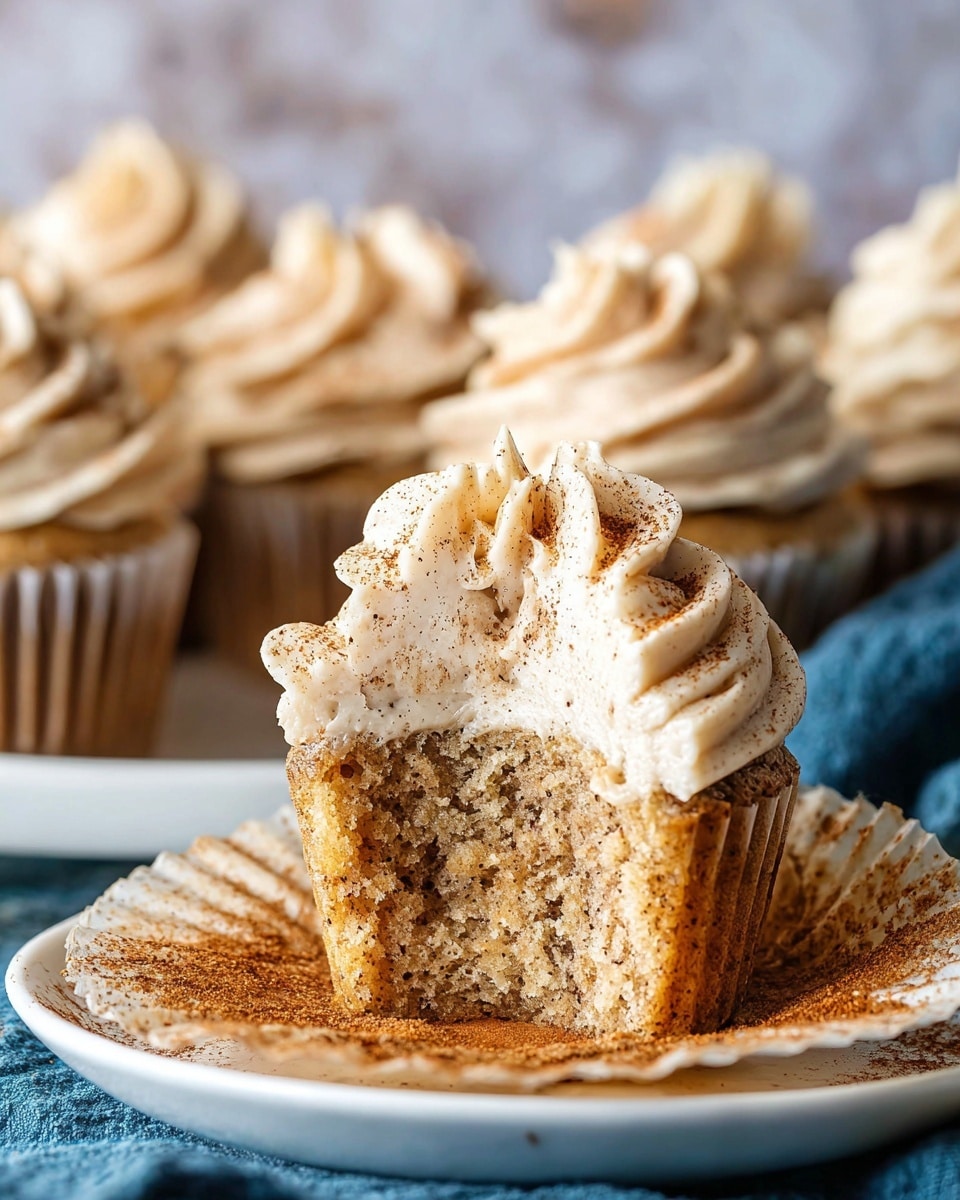 The image shows several cinnamon swirl cupcakes arranged on a white plate. Each cupcake has two visible layers: the base layer is a light brown cake with darker cinnamon swirls inside, and the top layer is a large swirl of creamy beige frosting sprinkled with cinnamon powder. The frosting sits high on each cupcake, forming soft peaks. Two cinnamon sticks lie beside the cupcakes on the plate. The plate rests on a folded light blue cloth, all placed on a white marbled surface with a dark blurred background. photo taken with an iphone --ar 4:5 --v 7