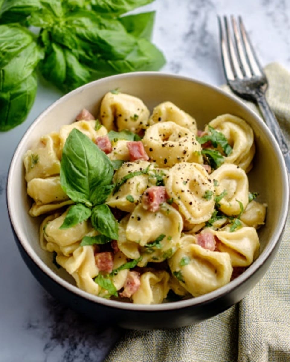 The image shows a white bowl filled with tortellini pasta mixed with small pieces of pink ham and green herbs, topped with fresh green basil leaves. The tortellini is creamy and slightly shiny, with a few black pepper specks visible on top. The bowl is placed on a white marbled surface, next to a light-colored cloth napkin and a silver fork. In the background, there are fresh green basil leaves in soft focus. Photo taken with an iphone --ar 4:5 --v 7