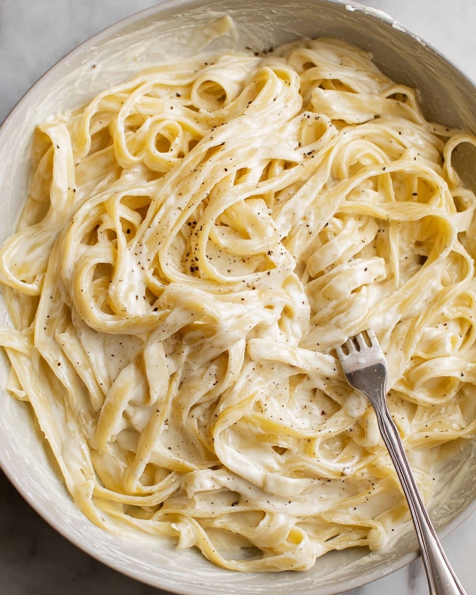 A close-up view of a white bowl filled with creamy pasta, showing a single layer of long, flat noodles coated in a smooth, pale cream sauce. The sauce appears thick and evenly covers the noodles, which are sprinkled lightly with small black pepper specks. A silver fork rests in the bowl, partially lifting some noodles, with the shiny, smooth texture of the metal contrasting the soft pasta. The bowl sits on a white marbled surface. photo taken with an iphone --ar 4:5 --v 7