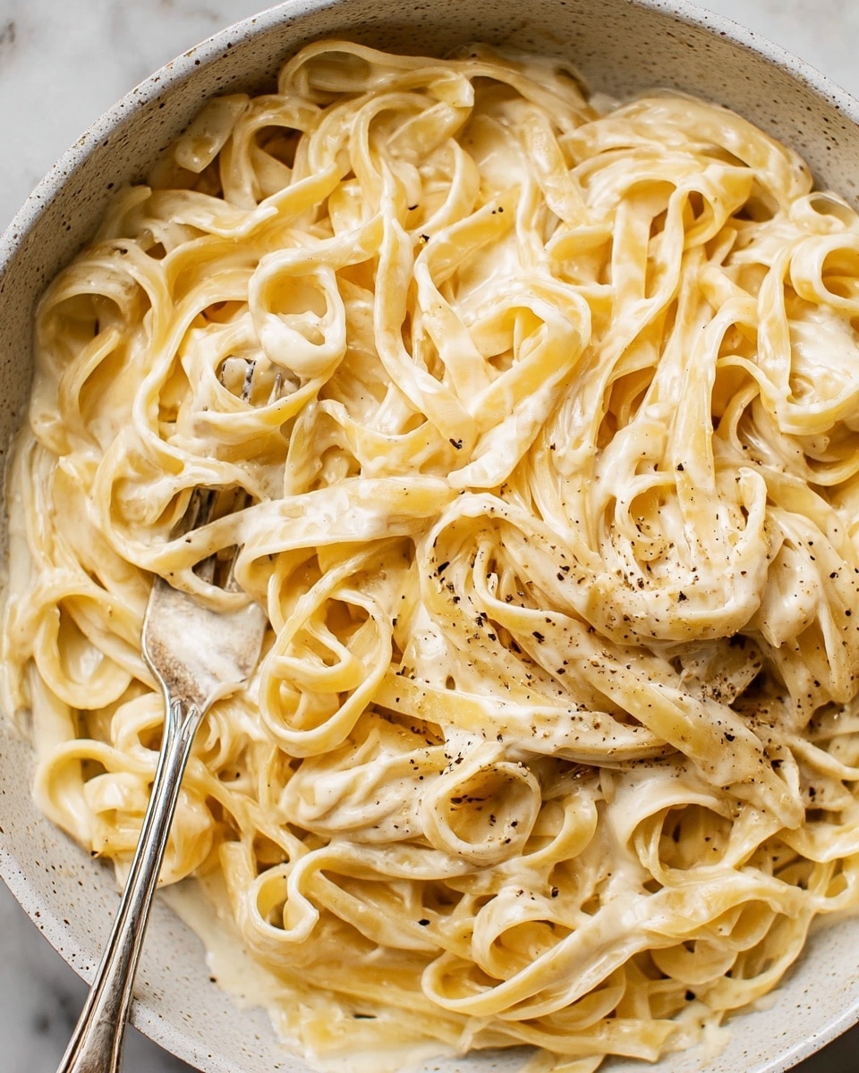 A close-up image of creamy fettuccine pasta in a white bowl with a slightly speckled texture, covered in a thick, smooth light beige sauce with black pepper sprinkled on top, showing glossy, well-coated noodles twisted and layered unevenly, with a silver fork resting inside the bowl. The bowl is placed on a white marbled texture. photo taken with an iphone --ar 4:5 --v 7