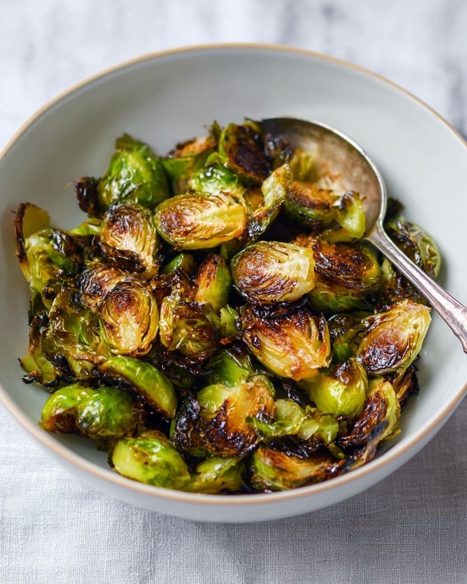 A white bowl filled with charred Brussels sprouts that have a mix of bright green, golden brown, and deep dark brown crispy edges. The Brussels sprouts are cut in halves and quarters, showing their layered texture with some leaves slightly loose and crispy. A silver spoon rests inside the bowl on the right side. The bowl sits on a dark blue cloth which contrasts with the white marbled background. photo taken with an iphone --ar 4:5 --v 7
