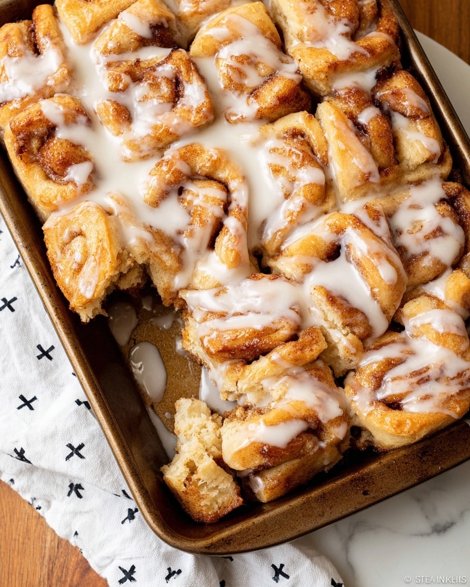 A close-up view of a metal baking tray filled with soft, golden brown cinnamon rolls that are torn into irregular chunky pieces, each piece showing layers of dough with a slightly crispy browned edge and a soft inside. The rolls are covered with thick white glaze that spreads unevenly across the surface, pooling in some places and dripping down the sides. The tray sits on a wooden table next to a white cloth with small black crosses, with all items placed on a white marbled textured surface. Photo taken with an iphone --ar 4:5 --v 7