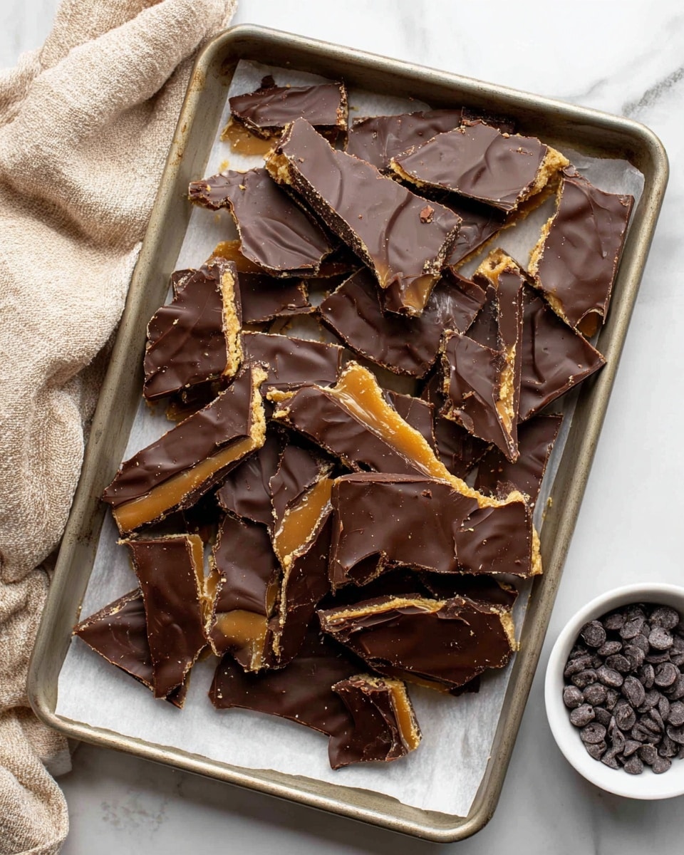 A white plate holds many pieces of cracked toffee bark arranged in a pile, each piece showing three layers: a rough golden brown brittle toffee base, a thin caramel layer in the middle, and a top layer of smooth, dark brown chocolate with visible swirling patterns and slight ridges; the plate is placed on a white marbled surface with green pine branches and frost-tipped pine cones around it, along with a beige cloth nearby. photo taken with an iphone --ar 4:5 --v 7