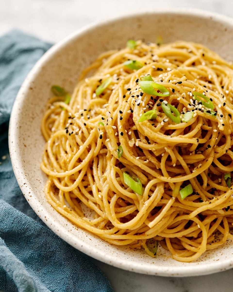 The image shows a white speckled bowl filled with a single layer of yellow spaghetti noodles that appear glossy and lightly coated with sauce, giving them a smooth texture. Scattered on top are small pieces of green sliced scallions and a sprinkling of black and white sesame seeds, adding color contrast and slight texture. The bowl sits on a white marbled surface with a blue cloth partially visible on the left side. photo taken with an iphone --ar 4:5 --v 7