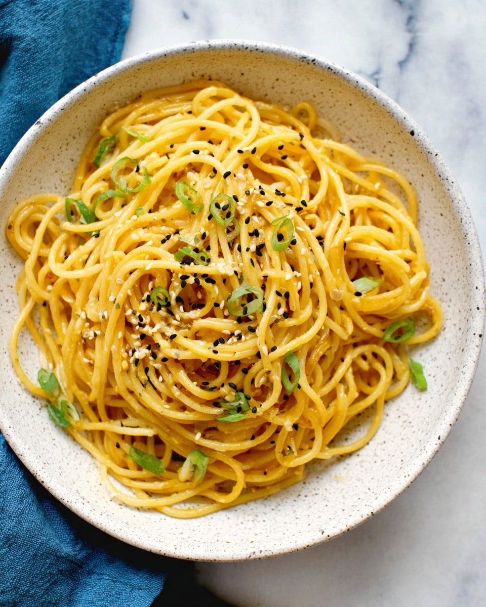 A white speckled bowl holds a mound of light brown spaghetti noodles twisted into a loose pile. The noodles are sprinkled with small black and white sesame seeds and garnished with thin slices of bright green scallions scattered on top. The bowl is placed on a white marbled surface, and there is a blue cloth partially visible on the left side. The image focuses closely on the noodles, showing their smooth, slightly shiny texture. photo taken with an iphone --ar 4:5 --v 7