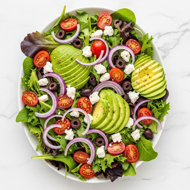 A fresh salad on a white oval plate with mixed green and purple lettuce leaves as the base layer, topped with bright red cherry tomato halves scattered evenly, sliced avocado pieces adding a soft green contrast, thin slices of purple onion spread across the salad, small black olives nestled throughout, and small white chunks of feta cheese sprinkled on top. The plate sits on a white marbled surface. Photo taken with an iphone --ar 4:5 --v 7