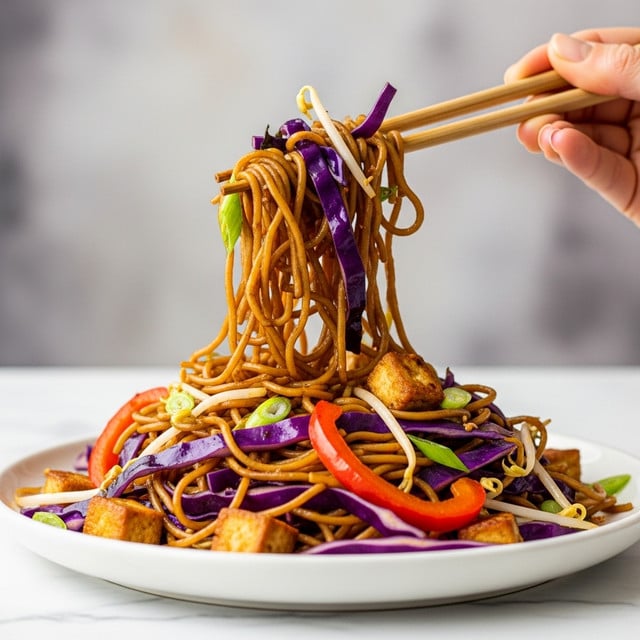 A close-up of a plate with a colorful noodle dish sits on a white marbled surface. The bottom layer is twisted brown noodles mixed with thin slices of bright orange carrots and purple cabbage. On top, there are darker browned cubes that look crispy, scattered around the noodles. Bright green chopped scallions and white sesame seeds are sprinkled across the dish. On the peak, light green bean sprouts add a fresh touch. In the background, there is a blurred blue cup. photo taken with an iphone --ar 4:5 --v 7