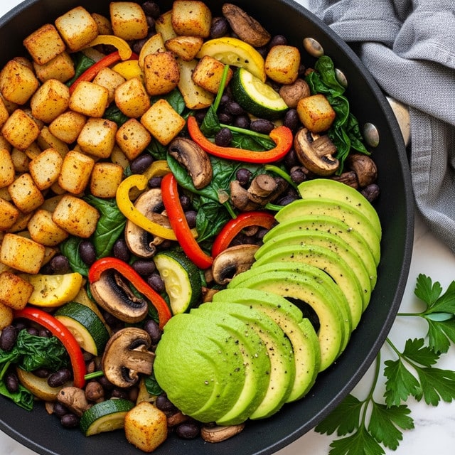 A close-up view of a black skillet filled with a colorful mix of cooked vegetables and cubed potatoes. The dish has three main layers: golden-brown crispy potato cubes on top and scattered throughout, bright yellow and green zucchini pieces, thin red bell pepper strips, dark green spinach leaves, and browned mushroom slices. Black beans are mixed in, adding dark contrast in small clusters. On the right side, a fan of light green avocado slices with slight black pepper specks rests on the vegetables. Fresh green parsley sprigs are placed around the skillet on a white marbled surface, with a gray cloth napkin in the upper right corner. photo taken with an iphone --ar 4:5 --v 7