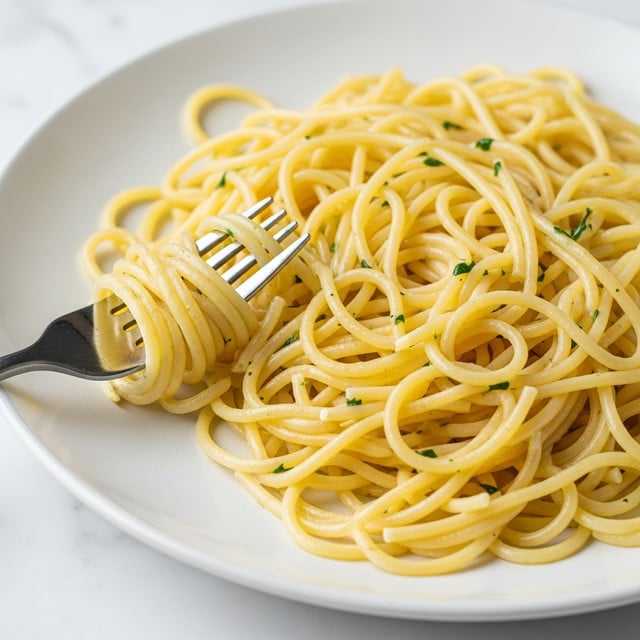 A close-up view of a plate with plain spaghetti pasta. The pasta is light yellow with a slight shine from oil and small bits of green herbs scattered throughout. The spaghetti is loosely tangled, with some strands wrapped around the end of a fork placed on the left side in the plate. The plate is white and round, set on a white marbled surface. photo taken with an iphone --ar 4:5 --v 7