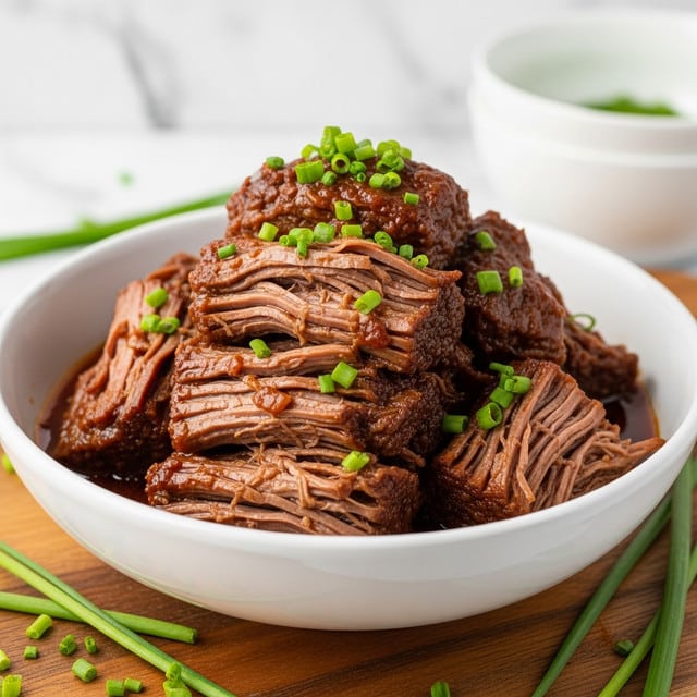 The dish shows a white bowl filled with braised beef chunks in a rich, dark reddish-brown sauce. The beef is shredded and layered on top of each other, soaked in the glossy sauce with visible textures of tender meat fibers. Bright green chopped chives are sprinkled on top and around the bowl, adding a contrast of color. The bowl is placed on a wooden board with some chives scattered outside. The background has a white marbled texture surface with a white bowl blurred in the back. Photo taken with an iphone --ar 4:5 --v 7