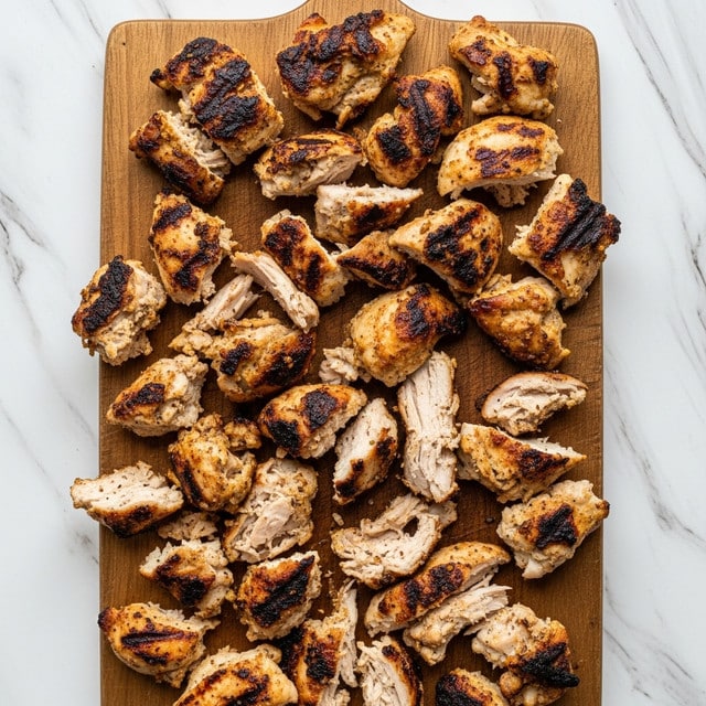 The image shows a wooden cutting board placed on a white marbled texture background with many pieces of grilled chicken spread across it. The chicken pieces vary in size and shape, with a mix of light golden brown and darker charred areas indicating a grilled texture. The chicken looks shredded and torn, showing a mix of white meat and slightly darker sections from the grill marks; some parts have a crispy crust while others appear tender. There are no other food items or utensils visible in the image. Photo taken with an iphone --ar 4:5 --v 7