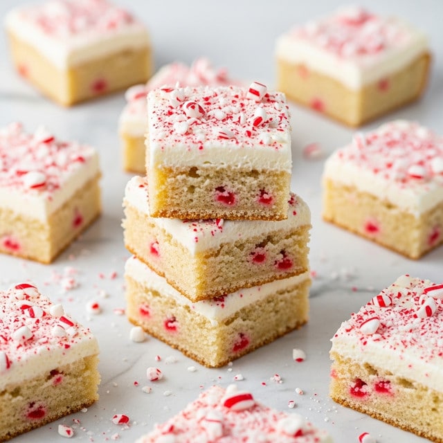 The image shows square dessert bars with three visible layers. The bottom layer is a light brown, crumbly base. Above that is a thin, slightly translucent red layer, suggesting a jelly or jam filling. The top layer is a thick, creamy white frosting sprinkled with crushed red and white peppermint pieces, giving a festive look. The bars are placed on a white marbled texture with scattered peppermint crumbs around them. photo taken with an iphone --ar 4:5 --v 7