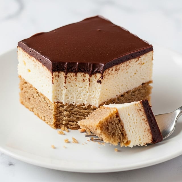 A close-up image of a square dessert piece on a white plate with three visible layers: the bottom layer is a light brown cake with a crumbly texture, the middle layer is a thick pale cream, and the top layer is a shiny, smooth dark chocolate ganache. A bite has been taken from the square, revealing the creamy interior and crumbly cake, with a fork holding a small piece of the dessert in front of the plate. The background is a white marbled texture. photo taken with an iphone --ar 4:5 --v 7