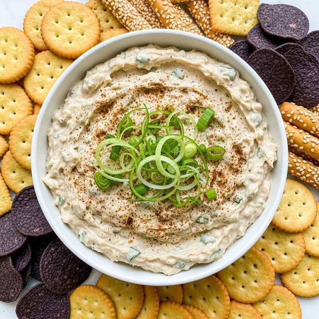 A close-up of a white bowl filled with creamy, light beige dip mixed with green bits and topped with thin, curly slices of green onion and a sprinkle of brown seasoning. The dip has a soft, slightly chunky texture. Around the bowl, there are various crackers including round golden crackers, square light brown crackers, sesame-topped breadsticks, and dark purple round chips, all placed on a white marbled surface. photo taken with an iphone --ar 4:5 --v 7