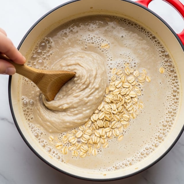 A close-up of a pot with a creamy beige mixture of oats and liquid simmering, showing small bubbles on the surface and chunks of oats floating halfway through the mixture, a wooden spoon stirring the thick texture from the left side held by a woman's hand, the pot has a white inside with a red outer edge, all set against a white marbled texture background. photo taken with an iphone --ar 4:5 --v 7