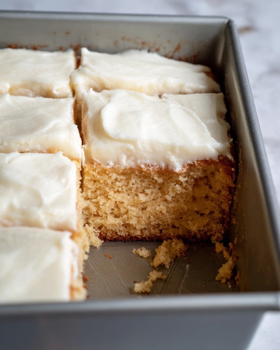 A close-up top view of a sheet cake cut into nine squares inside a baking pan. The cake has two visible layers: a moist, dense cake base in brown, and a thick, smooth, creamy white frosting layer on top. The frosting has a soft texture with slight waves and some small air bubbles. The pan edges are dark gray, and the cake is set on a white marbled texture with a striped cloth partially visible on the side. photo taken with an iphone --ar 4:5 --v 7