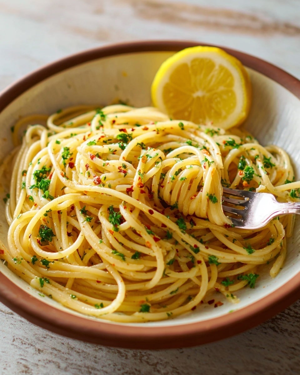 The image shows a bowl of spaghetti with two main layers. The base layer consists of twisted, smooth, light yellow spaghetti noodles, cooked and lightly coated with oil. On top, there are small green parsley flakes scattered evenly across the pasta and sprinkled red chili flakes for a slight hint of color. A silver fork is inserted into the spaghetti on the right side, lifting some noodles. Behind the pasta in the bowl, there is a bright yellow lemon wedge placed upright, adding a fresh touch. The round bowl is white with a brown rim, placed on a white marbled texture surface. Photo taken with an iphone --ar 4:5 --v 7