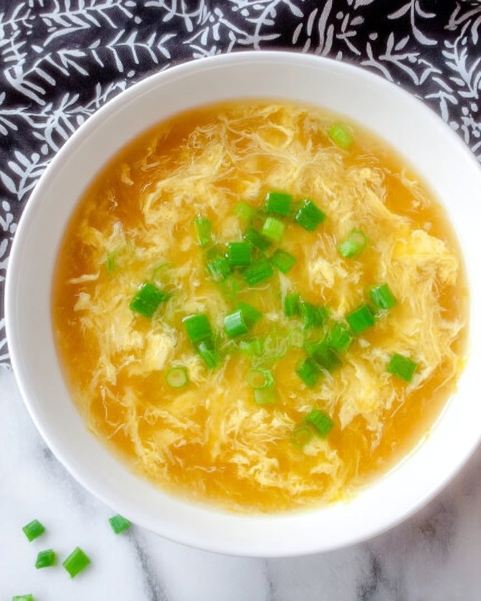 A close-up view of a white bowl filled with golden yellow egg drop soup, showing soft, wispy egg ribbons floating in the clear broth. Bright green chopped scallions are scattered on top, adding fresh color contrast. A white spoon rests inside the bowl, lifting some soup and eggs. The bowl is set on a white marbled surface with a blurred background showing a white egg carton. photo taken with an iphone --ar 4:5 --v 7