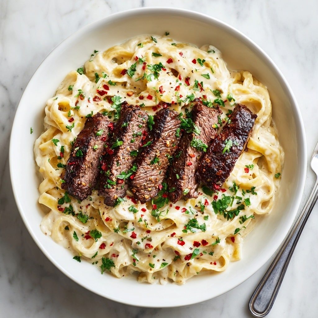 A white bowl filled with creamy fettuccine pasta coated in a smooth, off-white sauce, with visible herbs mixed in, creating a rich texture. On top, there are five pieces of grilled steak, browned with a slight char and grill marks, arranged in a neat line across the pasta. The dish is sprinkled with finely chopped green parsley and small red chili flakes scattered unevenly for a touch of color. The background is a white marbled texture. photo taken with an iphone --ar 4:5 --v 7