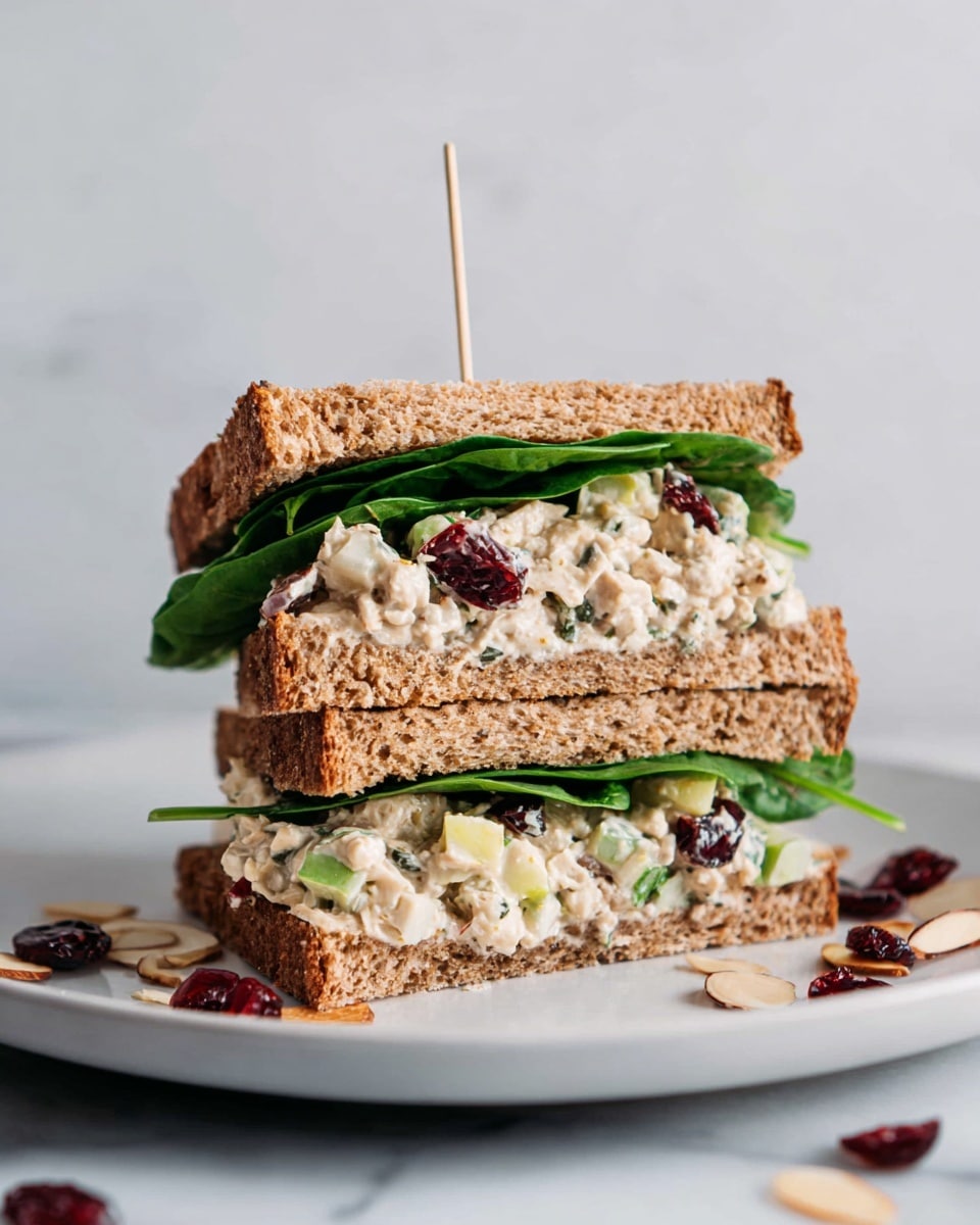 The image shows a sandwich cut in half and stacked, held together by a toothpick on top. It has three main layers: two slices of brown multigrain bread on the top and bottom with visible seeds and a rough texture, the middle layer is a creamy chicken salad mixed with small pieces of green celery, white apple chunks, and dark red dried cranberries, and fresh green spinach leaves are layered between the bread and chicken salad. The sandwich is placed on a white plate, set on a white marbled surface with some sliced almonds and dried cranberries scattered nearby. photo taken with an iphone --ar 4:5 --v 7