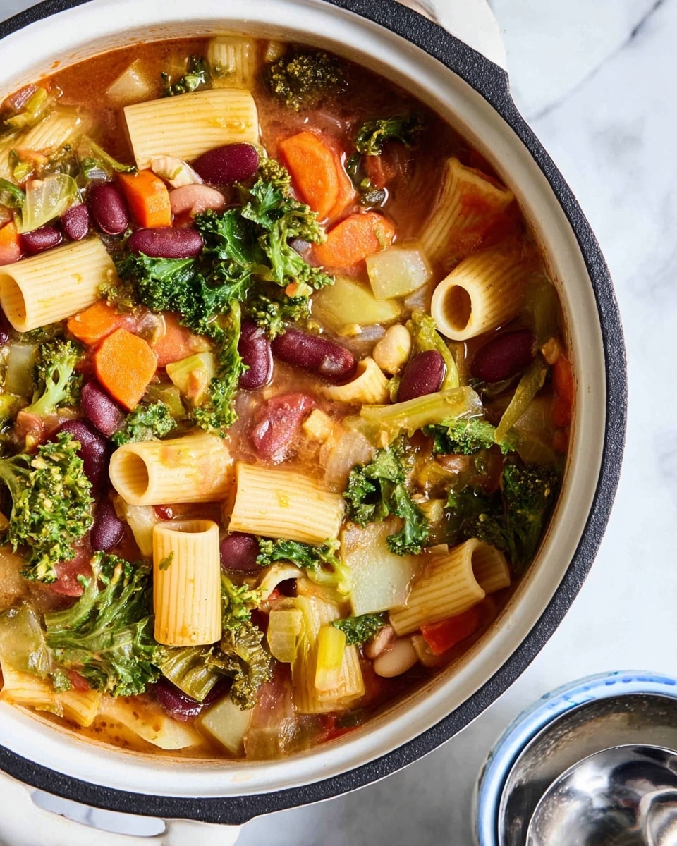The image shows a close-up of a white enamel cooking pot with a black rim, filled with a colorful vegetable soup. The soup has large tube-shaped pasta in pale yellow, dark red kidney beans, bright orange carrot slices, green broccoli florets, leafy dark green kale, and small chunks of light beige potato, all immersed in a slightly thick light brown broth. Some pieces of diced onion and celery are scattered throughout, adding texture and lightness. The pot sits on a white marbled surface, and a white bowl with a blue rim is partially visible at the bottom right corner next to a shiny silver ladle. Photo taken with an iphone --ar 4:5 --v 7