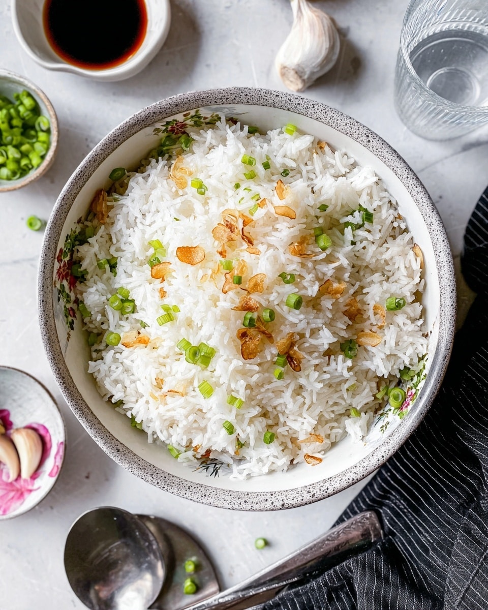 A bowl filled with steamed white rice topped with small pieces of golden toasted garlic and bright green chopped spring onions, giving a mix of white, light brown, and green colors. The bowl is mainly gray with floral patterns inside visible on the sides. Surrounding the bowl is a glass of water with ice, a small white dish with dark sauce and a metal spoon, a white bowl holding more chopped green onions, garlic cloves placed on a white marbled surface, and folded cloth napkins in black and white patterns. photo taken with an iphone --ar 4:5 --v 7