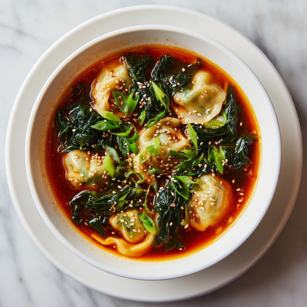 A white bowl is filled with a spicy broth that has a rich reddish-brown color. Floating in the broth are several light beige dumplings with smooth, slightly translucent wrappers. Bright green chopped scallions and wilted dark green leafy vegetables are scattered around the dumplings. White sesame seeds sprinkle the surface, adding a textured look. The bowl sits on a white plate, placed on a white marbled texture. Photo taken with an iphone --ar 4:5 --v 7