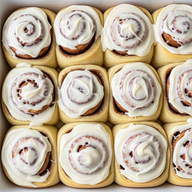 A white baking dish filled with ten cinnamon rolls arranged close to each other in two rows. Each cinnamon roll has a visible spiral shape with a light golden-brown bread layer and a thick, creamy white icing spread unevenly over the top, covering most of the rolls except slight hints of the cinnamon spiral showing through. The texture of the icing looks smooth and glossy, slightly melted and dripping down the sides of some rolls. The background is a white marbled texture. Photo taken with an iphone --ar 4:5 --v 7
