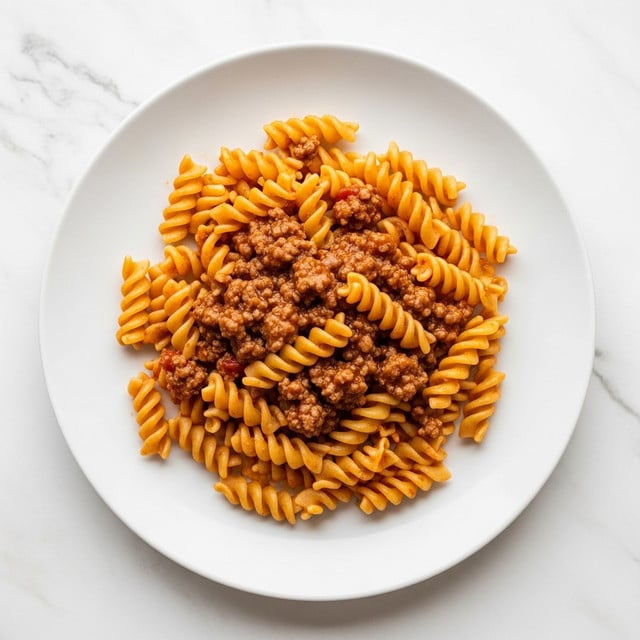 A round white plate holds a single layer of spiral-shaped rotini pasta mixed with a thick, rich meat sauce. The sauce is brown with visible small chunks of ground meat and bits of red tomato, evenly coating the yellow-orange pasta spirals. The plate sits on a white marbled surface, and the photo is taken from above, showing the pasta pile centered neatly on the plate. photo taken with an iphone --ar 4:5 --v 7
