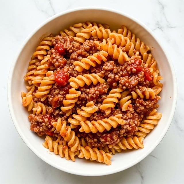 A white bowl filled with rotini pasta mixed with a thick, rich meat sauce. The sauce has a deep reddish-brown color with visible small chunks of ground meat and bits of red tomato, coating and filling the pasta spirals evenly. The pasta is spread in a loose mound, showing its firm texture. The bowl sits on a white marbled surface, with soft natural light highlighting the glossy sauce and pasta curves. photo taken with an iphone --ar 4:5 --v 7