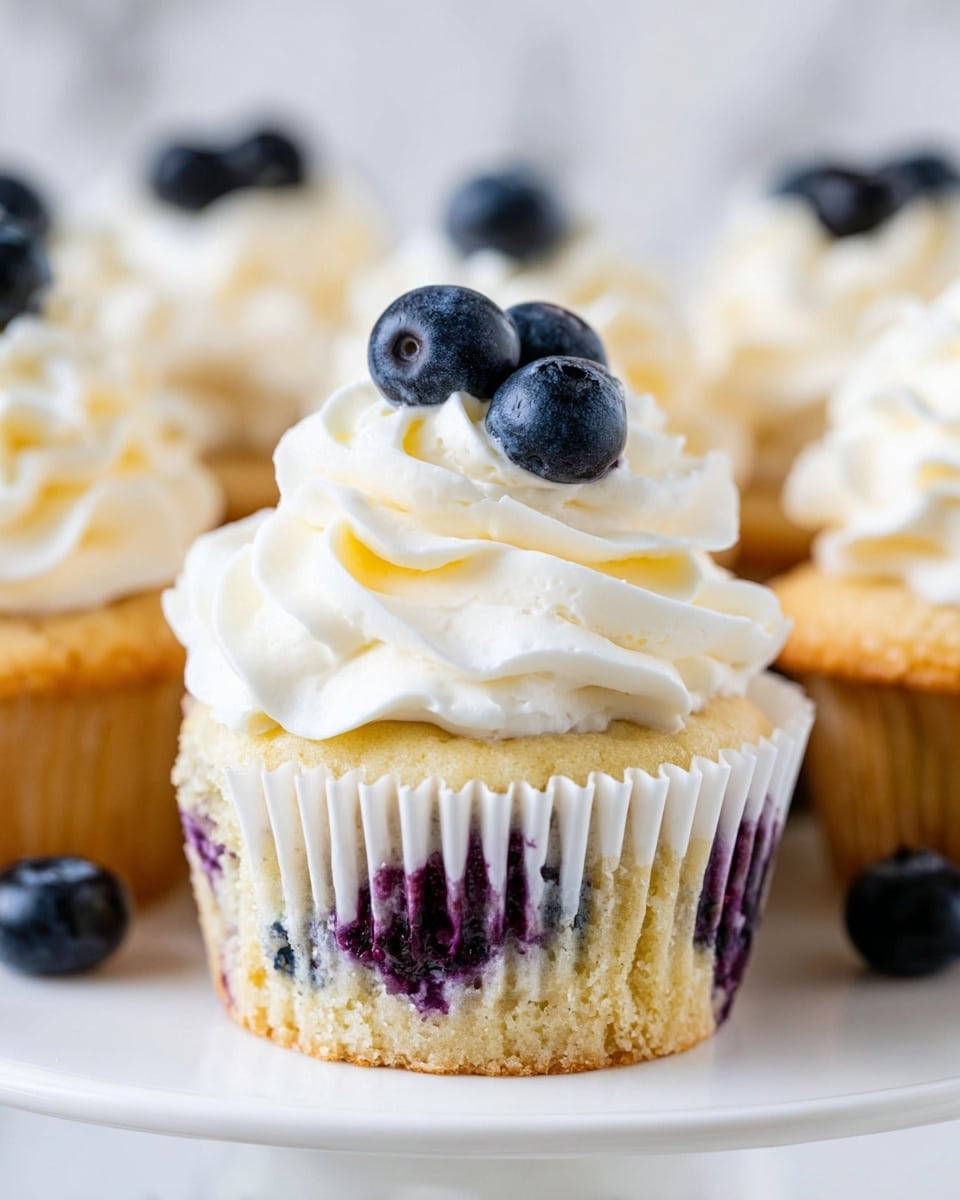 The image shows several blueberry cupcakes arranged on a white marbled surface. Each cupcake has a yellowish base dotted with purple blueberry spots visible through the white paper wrappers. On top, there is a thick swirl of smooth white frosting, piped in a rounded spiral with soft peaks. Each frosting swirl is decorated with two fresh blueberries placed near the center. The focus is sharp on the cupcake in front, with others softly blurred in the background, creating a fresh and inviting look. photo taken with an iphone --ar 4:5 --v 7