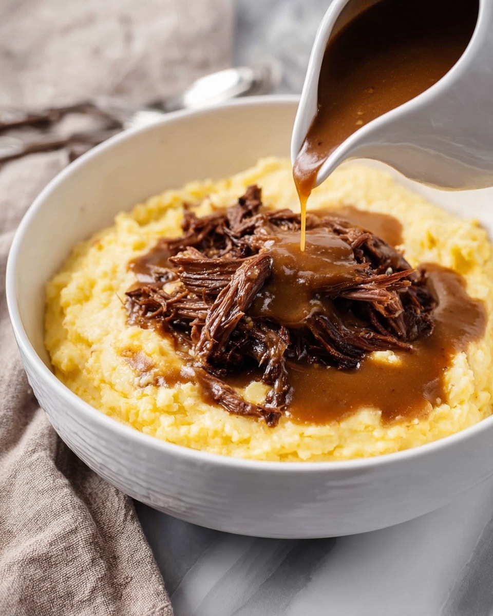 A white bowl filled with a base layer of creamy, light yellow mashed potatoes that have a smooth texture with slight lumps. On top, there is a thick, dark brown gravy spread around the edges and partly covering the potatoes. At the center, there is a pile of shredded, tender beef pieces with a rich, moist look, soaked in the same gravy. A spoon rests on the right side inside the bowl. The bowl sits on a white marbled textured surface with some sprigs of green herbs nearby. photo taken with an iphone --ar 4:5 --v 7