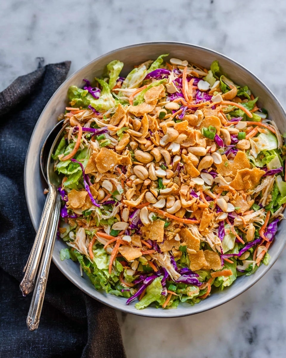 A large black bowl filled with a colorful salad sits on a white marbled surface, with a folded blue cloth nearby. The salad has three main layers: the bottom layer is light green lettuce leaves, the middle layer shows thin purple cabbage strips and orange carrot shreds mixed with pieces of cooked chicken, and the top layer is made of golden crispy fried noodles and sliced almonds scattered all over. Two silver salad tongs rest inside the bowl at the back. The background is softly blurred with warm, earthy brown tones. Photo taken with an iphone --ar 4:5 --v 7